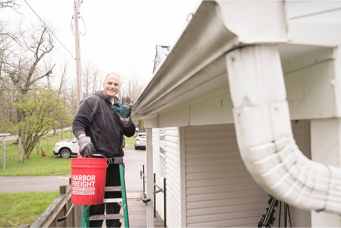 BBW employee on a ladder cleaning out a gutter and waving to the camera.