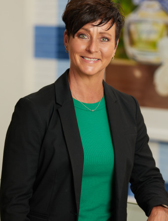Renee Barton-Robare is posed, smiling. She is standing in front of a gingham background, wearing a black blazer.